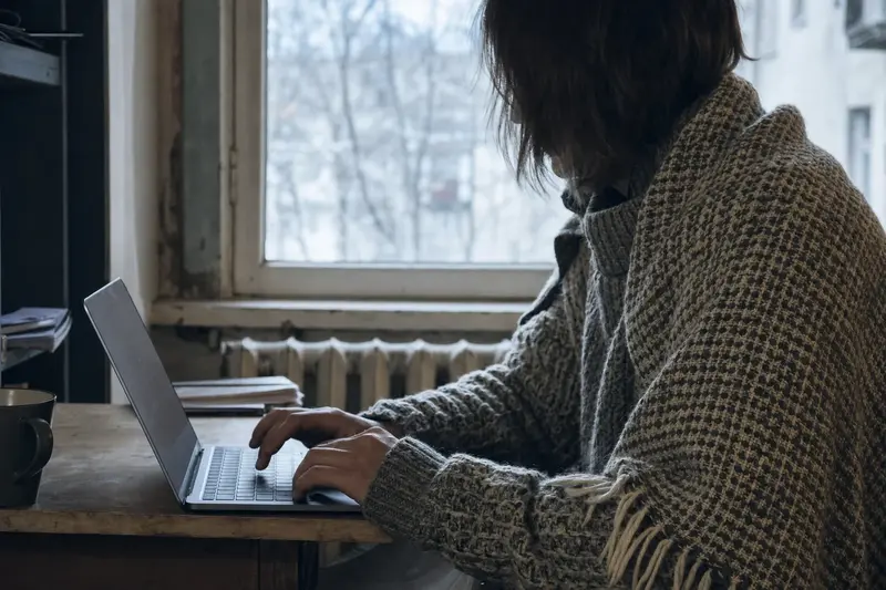 Človek zabalený v deke pracuje na notebooku pri okne s radiátorom