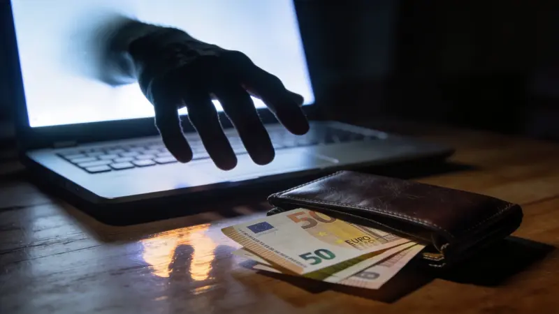 Tmavá ruka siahajúca z obrazovky notebooku po peňaženke s eurami